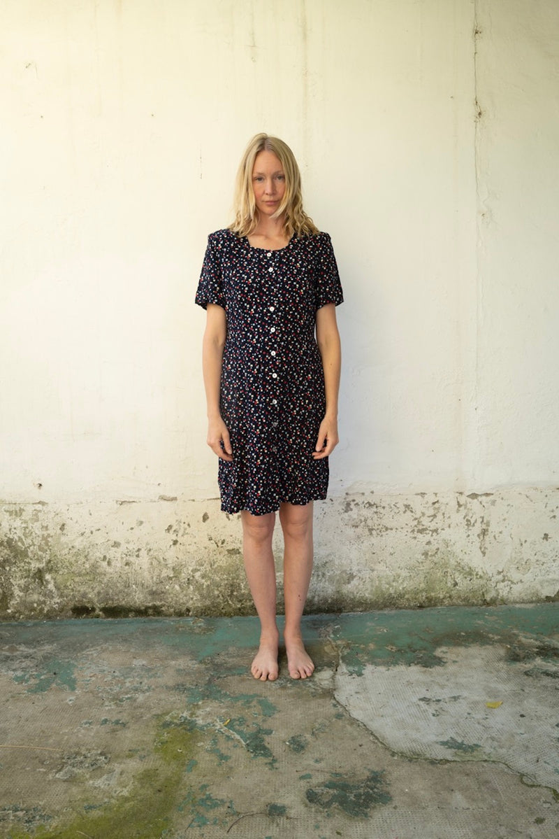 Woman wearing a black patterned dress standing against a white wall.
